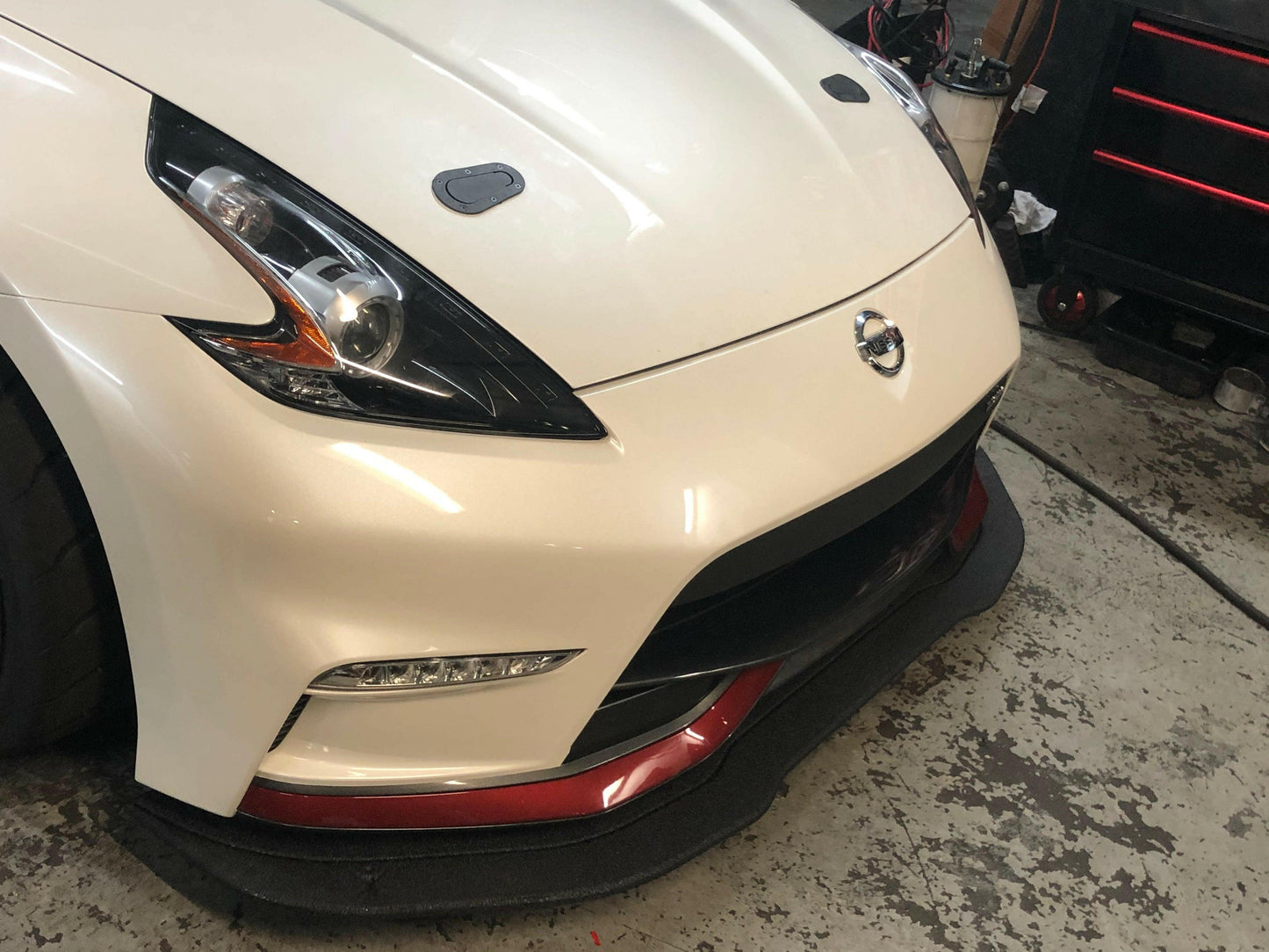 Close-up image of the front corner of a white Nissan 370z featuring a sleek headlight design, a black front lip with red accents, and a Nissan emblem on the grille. The 9livesracing 370z Rocket Nose v2 Splitter ’08-20 Z34 enhances downforce for superior performance. The car is parked in what appears to be a garage or workshop with tools visible in the background.