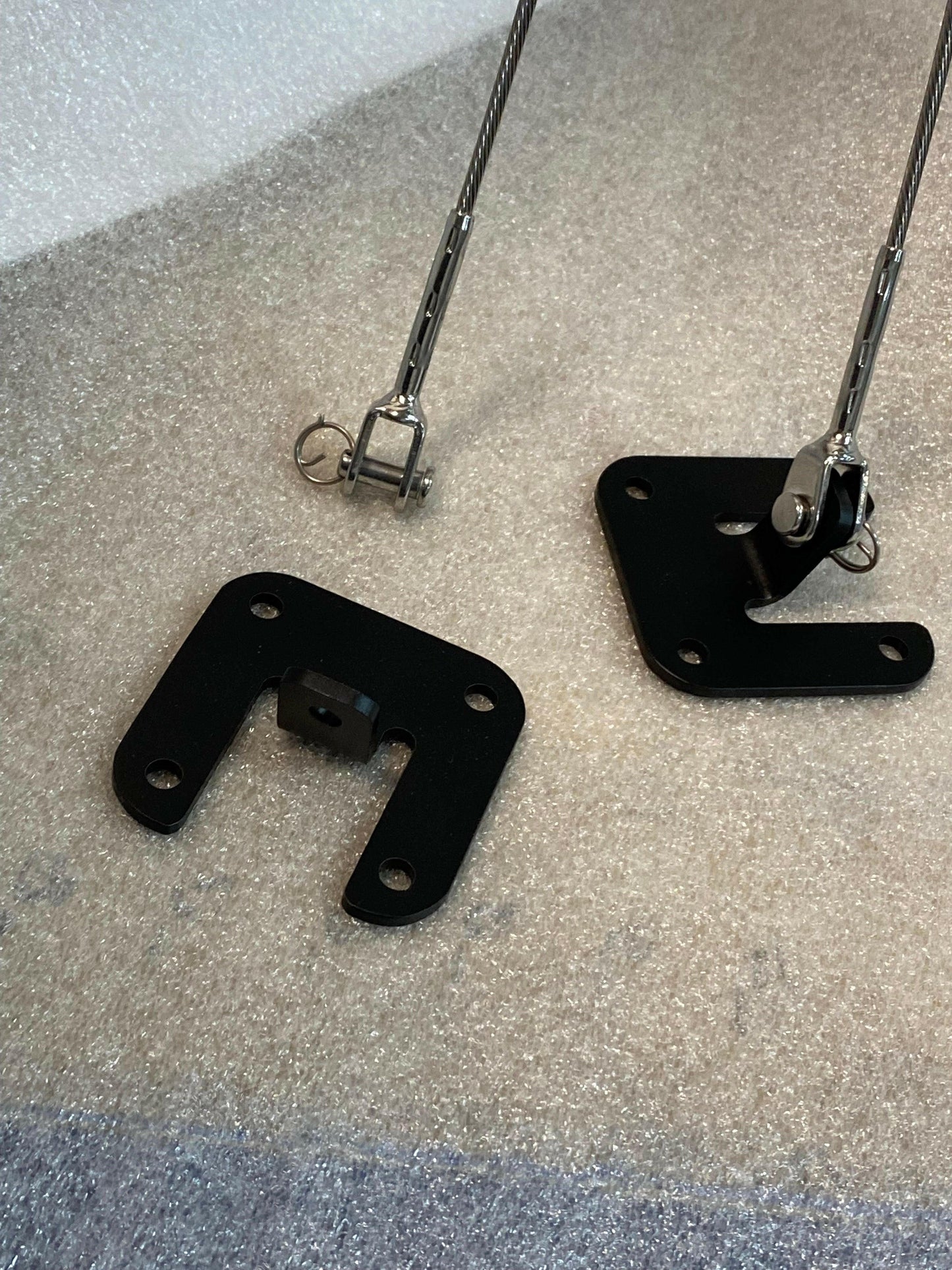 Two black metal brackets with multiple holes and a small metal pin inserted in one of the holes are placed on a white, textured surface. These Nine Lives Racing Splitter Side Supports have attached threaded rods with eyelets at the ends, indicating they are adjustable components perfect for racecar accessories.