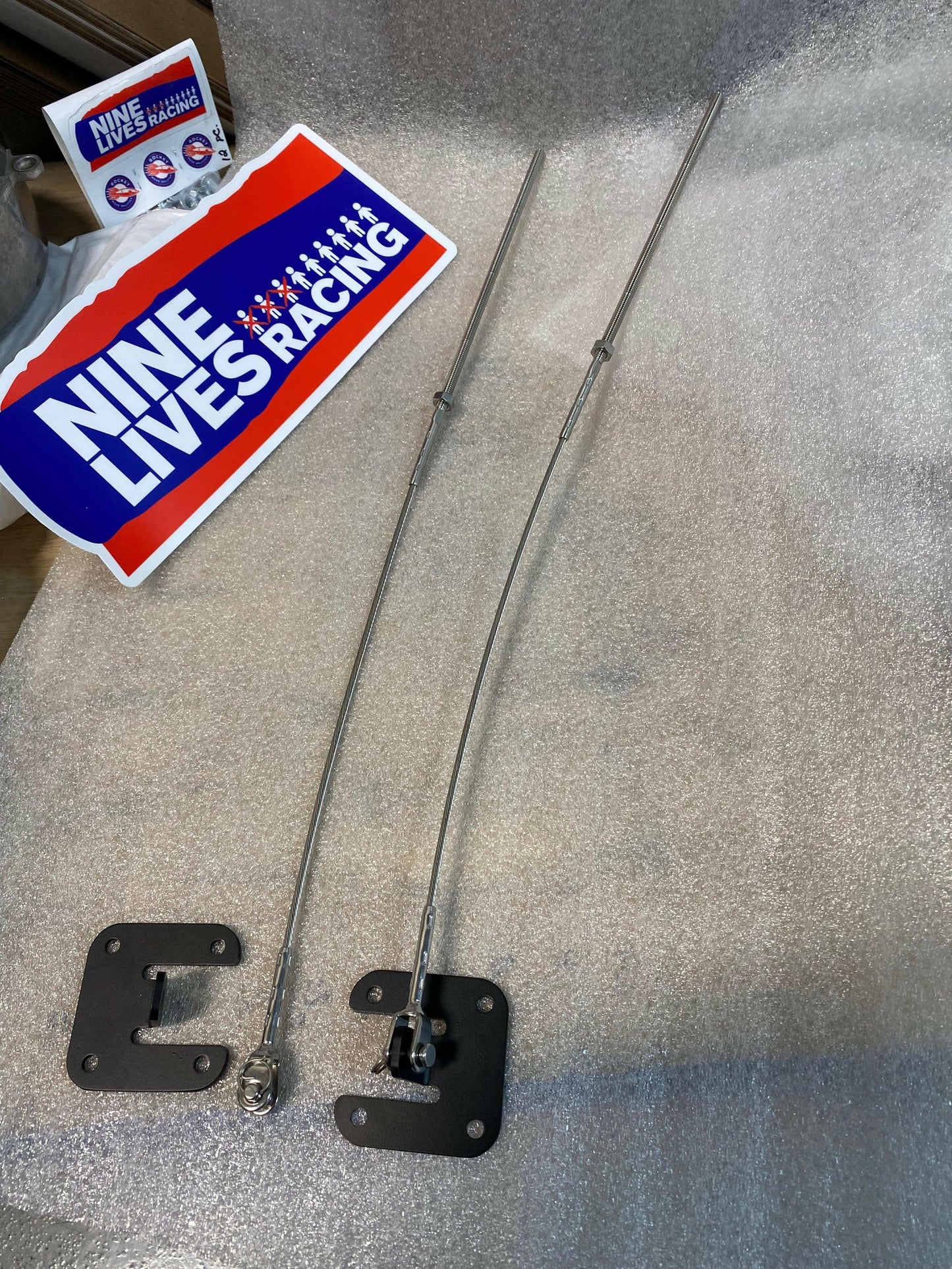 Two metal Splitter Side Supports with black brackets are laid out on a textured surface. Beside them is a sticker displaying "Nine Lives Racing" in large white letters on a blue background. This racecar accessory also features smaller text and several logo variations nearby.