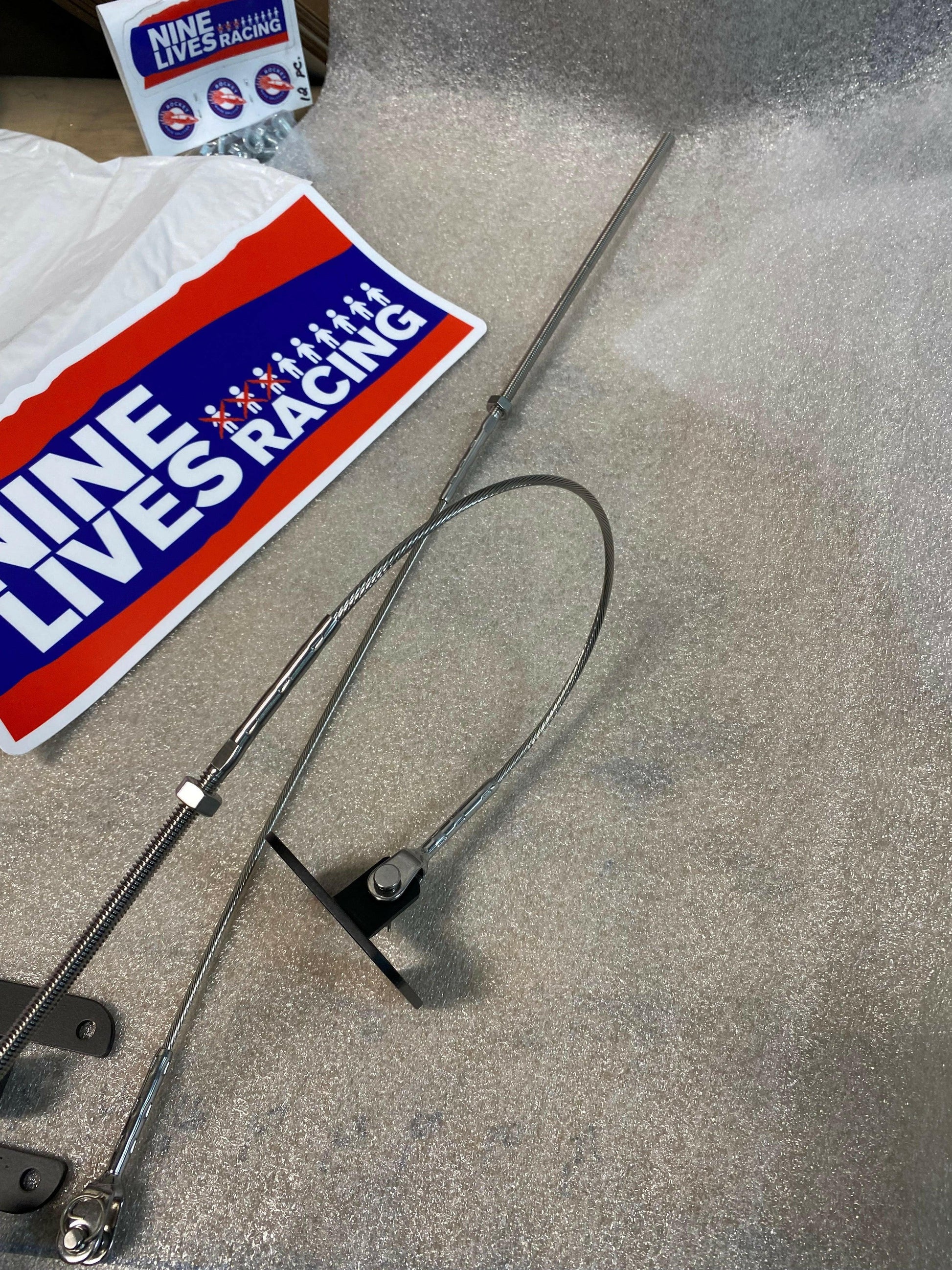 Close-up of a package containing Splitter Side Supports from Nine Lives Racing, featuring metal rods and stainless cables arranged on a textured foam surface. A Nine Lives Racing logo sticker is visible in the background, highlighting their premium racecar accessories.