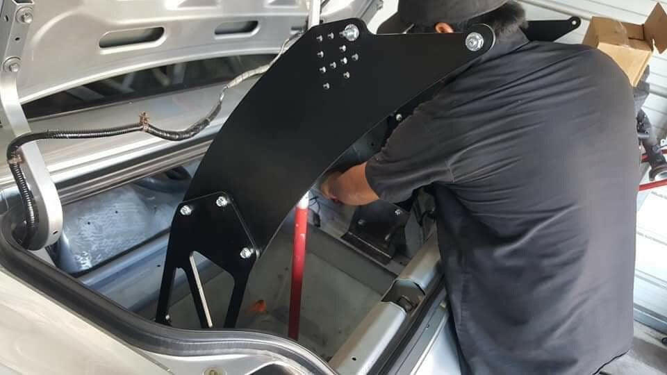 A person wearing a black shirt and cap installs the Honda S2000 Chassis Pylons - Pylons only with aluminum hardware from 9livesracing in the trunk of a vehicle. The trunk lid is open, revealing various tools and components.