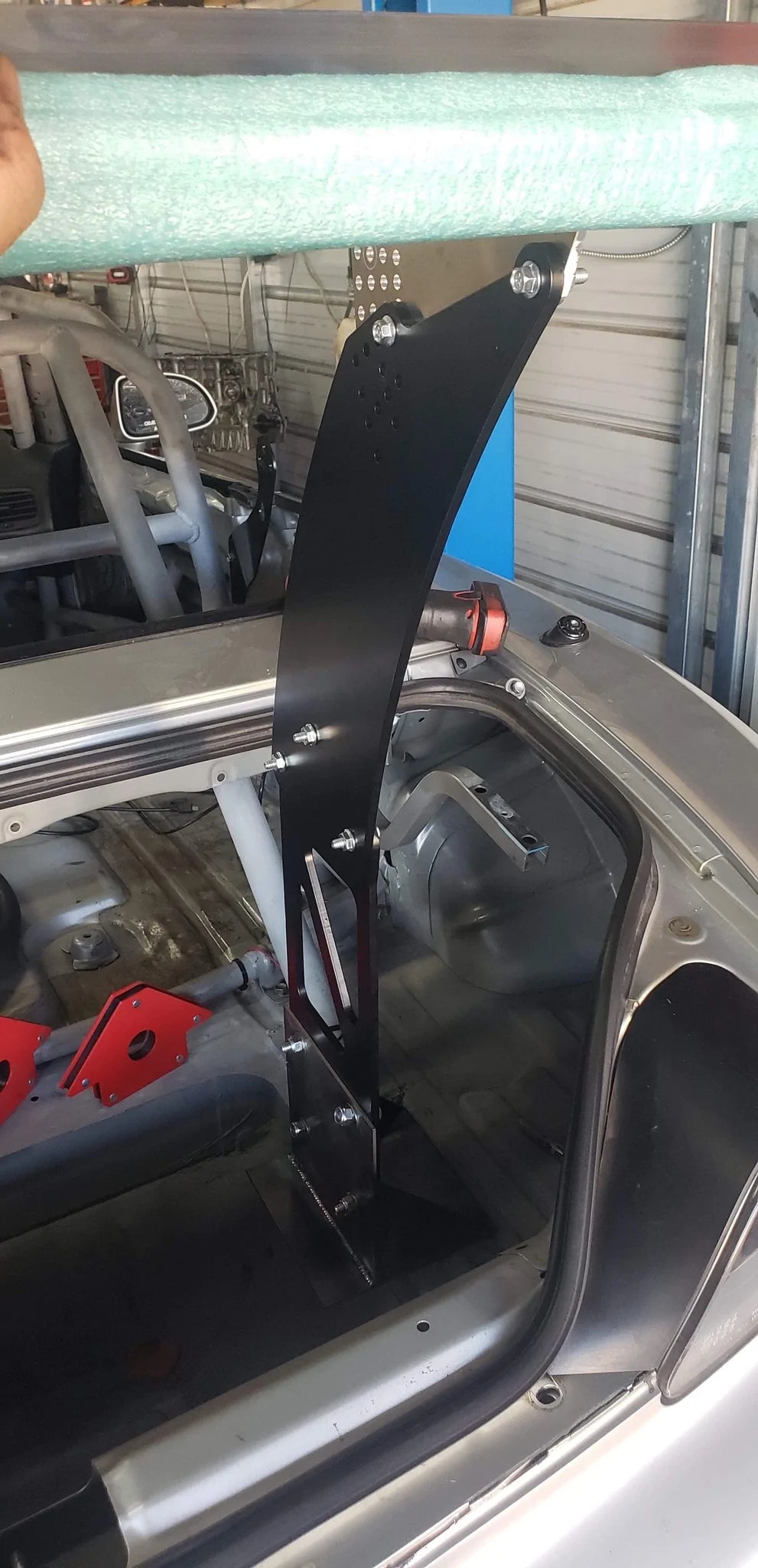 A black metal Honda S2000 Chassis Pylon by 9livesracing extends upward from the rear interior of a partially assembled car, secured with aluminum hardware. The pylon is anchored to the car’s body, and a hand is holding a padded green bar above it. The car's roll cage and other components are visible.