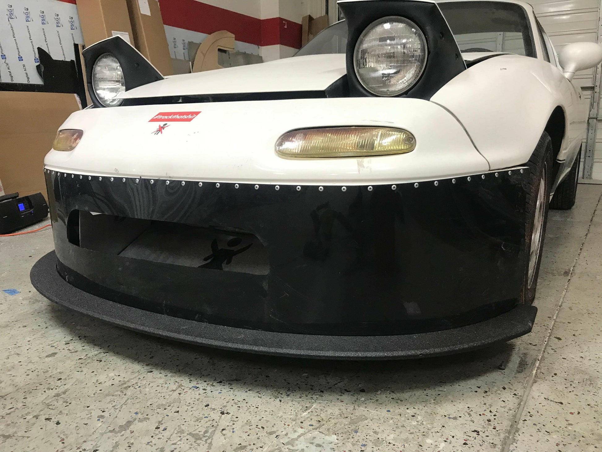 A white Mazda Miata with pop-up headlights and a large black front splitter from 9livesracing stands parked in a garage. The car, featuring a red emblem on the front, utilizes its Rocket Nose design for enhanced downforce. Various items are scattered in the background while the photo highlights the front bumper and splitter, which is designed for Mazda Miatas from 1990-05 NA/NB models.