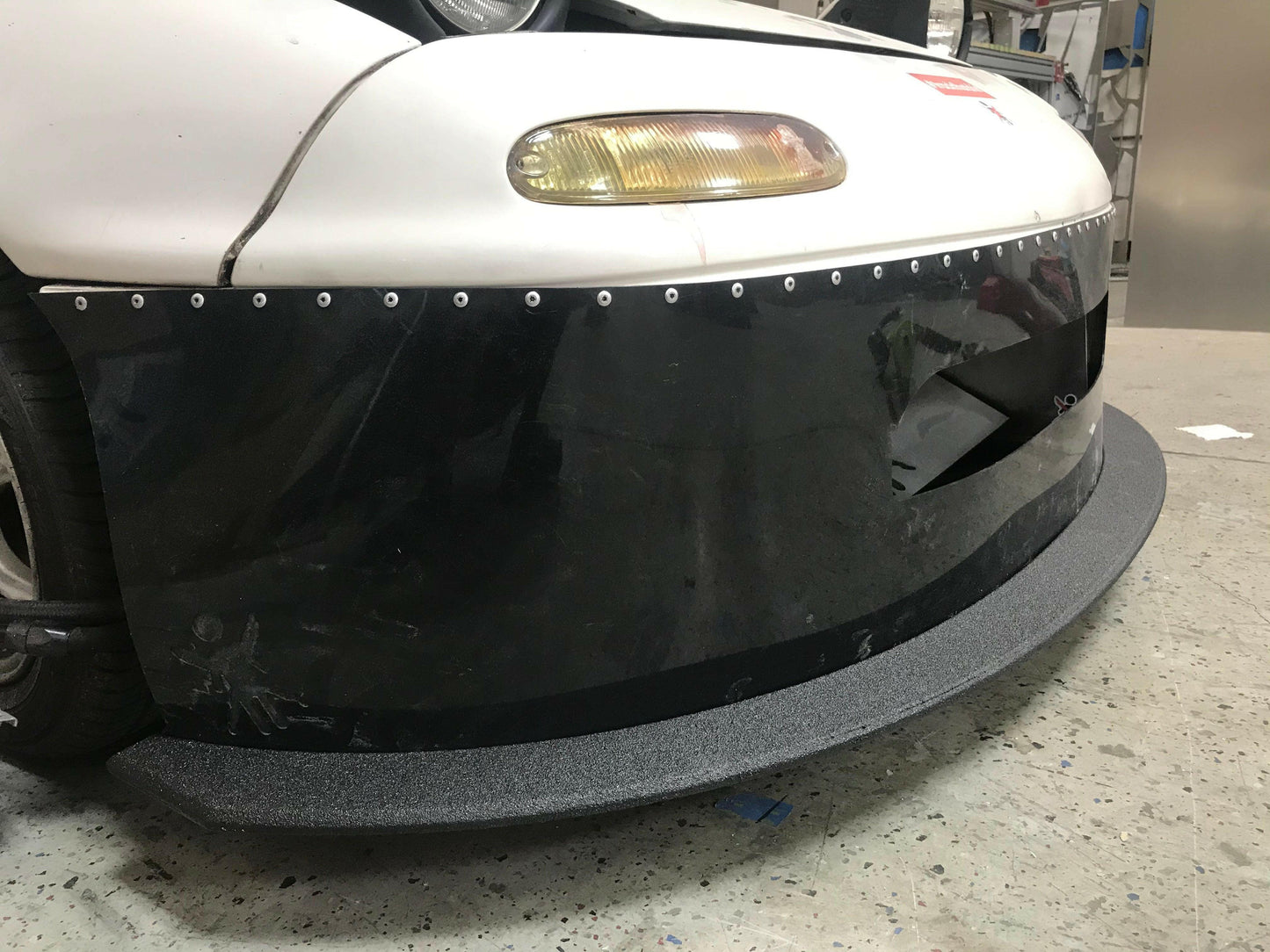 Close-up of the front end of a car featuring a white hood, yellow-tinted headlights, and a black front bumper with rivets and a textured gray Mazda Miata Splitter (1990-05 NA/NB) by 9livesracing. The car's aggressive Rocket Nose design enhances downforce, parked on a concrete floor in a garage or workshop with blurred tools and equipment in the background.