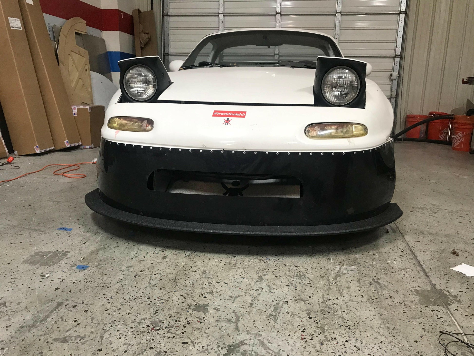 A white Mazda Miata, equipped with raised pop-up headlights and a sleek rocket nose, is parked in a garage. The car features a black front bumper and proudly displays a red emblem on the hood. Assorted items and equipment are visible in the background, enhancing its high-performance aura.