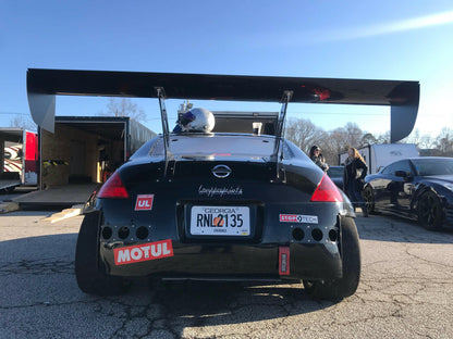 A black sports car with a massive 9livesracing pre-preg carbon wing and various decals, including Motul and StopTech, is parked on a paved area. The license plate reads "RNL7135" from Georgia. A helmet is resting on the rear of the car, and a secondary car is visible nearby.