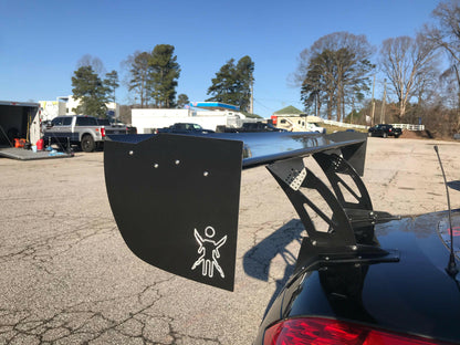 A black sports car, equipped with a large 9livesracing carbon wing, sits in a parking lot. The spoiler, designed to maximize down-force, prominently features the distinctive 9livesracing logo of a figure with spread arms. In the background, other parked vehicles, buildings, and tall trees stand under a clear blue sky.