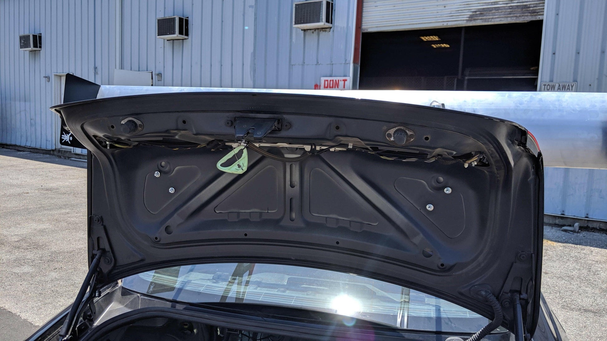 A car with its trunk open is parked in front of an industrial building. The interior of the trunk lid reveals metal reinforcements and wiring, while the 3-Series Big Wang kit ’04-13 E90/E92/E93 from 9livesracing, featuring extruded aluminum wings for added stability, is visible. The building's exterior consists of corrugated metal walls with several air conditioning units attached. Parts of another parked car can also be seen nearby.