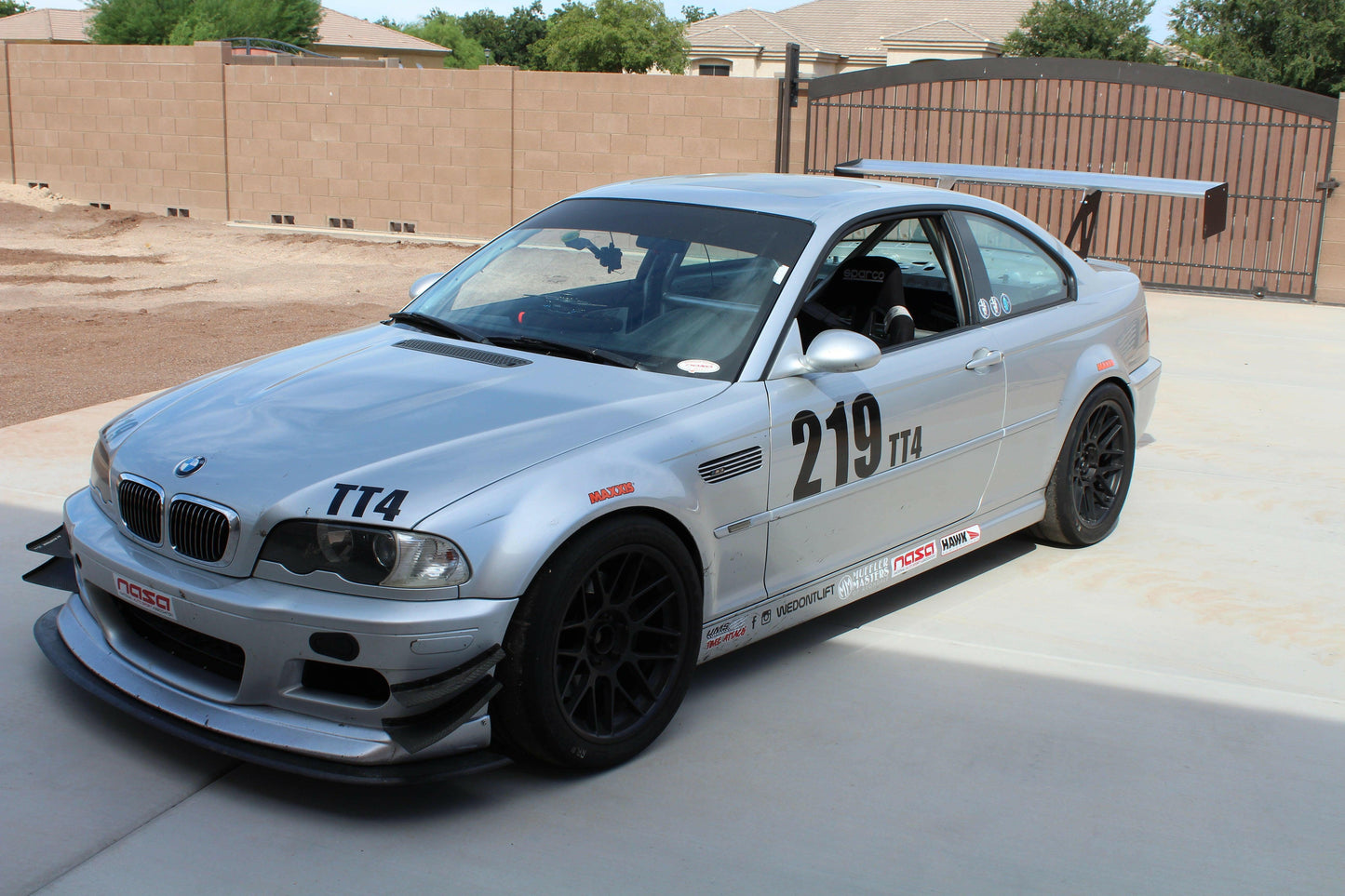 A silver BMW E46 M3 race car with black wheels, a 9livesracing 3-Series Big Wang Kit '97-06 E46 rear wing, and racing number 219 is parked on a driveway. The car showcases various sponsor decals and modifications, including front splitters and a roll cage. A fenced yard and neighboring houses can be seen in the background.