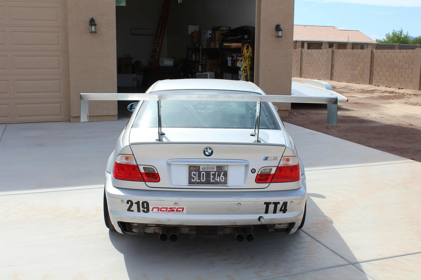 A silver BMW E46 race car is parked on a cement driveway. The car features an extruded aluminum 3-Series Big Wang Kit '97-06 E46 from 9livesracing, quad-exhaust, and a customized license plate reading "SLO E46." Racing decals and the number "219" are on the bumper. A garage with tools and equipment is in the background.