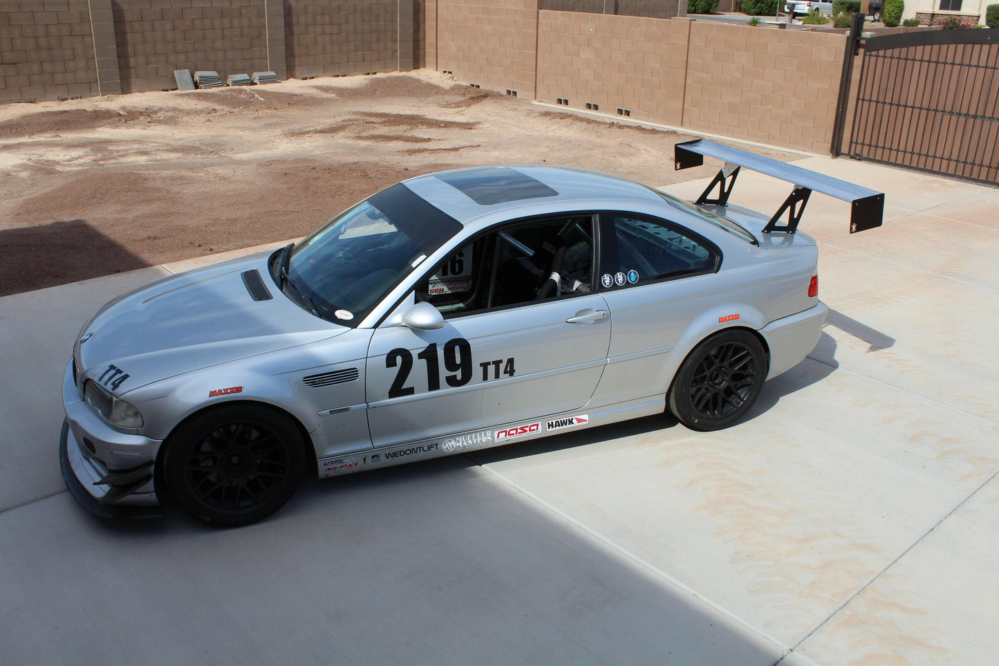 A silver race car, bearing the number 219 and TT4 on its side, is parked on a concrete driveway. The 3-Series Big Wang Kit '97-06 E46 model by 9livesracing boasts a large rear wing spoiler and black wheels. It is situated in an enclosed area with a brick wall and a metal gate, with racing decals adorning the sides of the car.