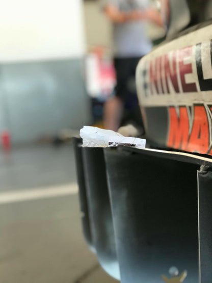 Close-up of the rear section of a vehicle, probably a car, highlighting the Miata Diffuser '90-05 NA/NB by 9livesracing with a small piece of crumpled white paper lodged in it. The background is out-of-focus, featuring people and some equipment.