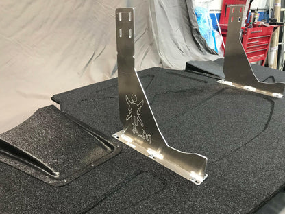 Two metal brackets from the Corvette Medium Downforce Kit ‘97-04 C5 by 9livesracing are installed on a textured black surface. The brackets feature small cutouts, with each one displaying an engraved figure with outstretched arms and the label "D-1." In the background, a workbench with red drawers and various tools can be seen, likely parts of this aerodynamic kit for the C5 Corvette.