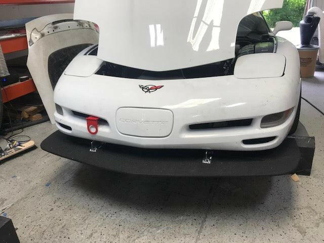 A white sports car with its hood open in a garage or workshop, showcasing 9livesracing Corvette Sturdy Boii Splitter Mounts (‘97-04 C5) and a red tow hook on the front bumper. The car, positioned on a black mat, distinctly bears the Corvette emblem with various tools and objects visible on either side of the image.