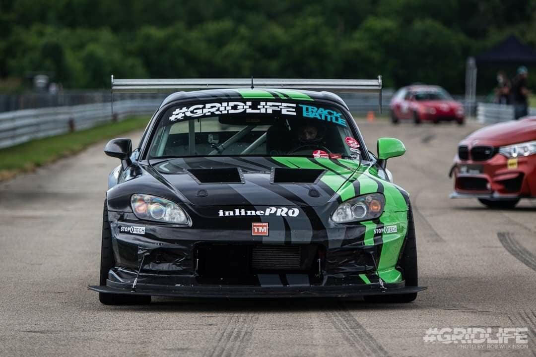 A black and green race car with "Gridlife" on the windshield, equipped with a sleek S2000 Rocket Nose Splitter for Voltex bumpers ’99-09 by 9livesracing, speeds down a racetrack. Another car can be seen in the background. The track, bordered by trees and safety barriers, exudes the atmosphere of a competitive racing event.