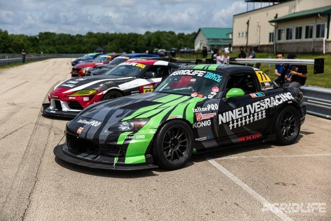 A lineup of colorful, modified race cars parked on an asphalt surface near a racetrack. The closest car, featuring a vibrant green and black design adorned with various sponsor logos including "9livesracing," sports an S2000 Rocket Nose Splitter for Voltex bumpers ’99-'09 with intricate CNC cut details. A building and trees are visible in the background.