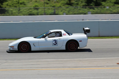 A white racing car with the number 3 on its side is speeding on a racetrack. Equipped with the Corvette Medium Downforce Kit ‘97-04 C5 from 9livesracing, this C5 Corvette boasts red rims and a large rear spoiler, creating significant downforce. An American flag sticker near the driver's door adds flair. The background features barriers, grassy areas, and trees.