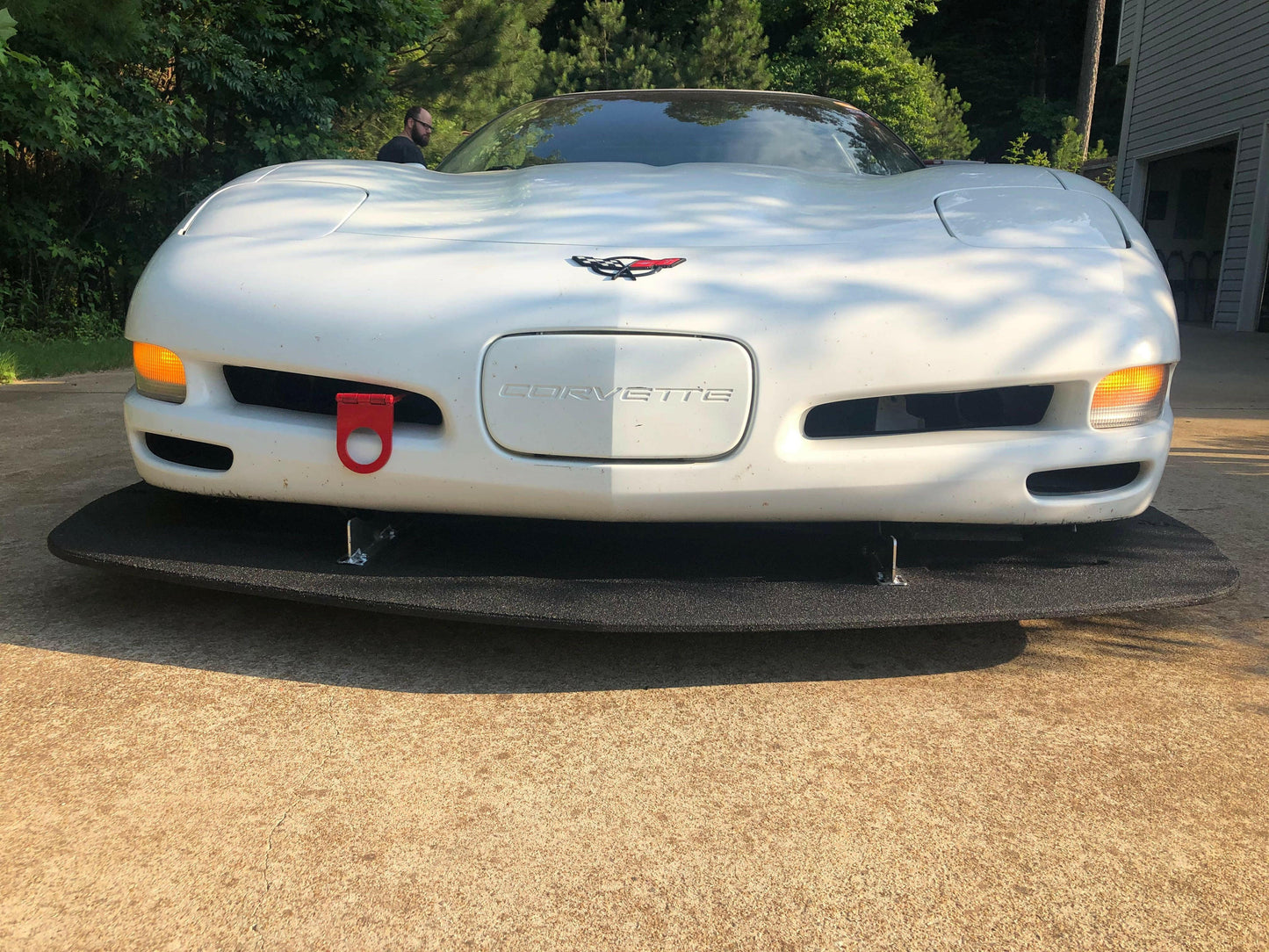 A white Corvette is parked on a driveway, featuring 9livesracing's Corvette Sturdy Boii Splitter Mounts '97-04 C5 for added downforce. The front bumper boasts a red tow hook on the left side, the iconic Corvette emblem in the center, and yellow turn signal lights on both sides. In the background, a house stands to the right and trees to the left.