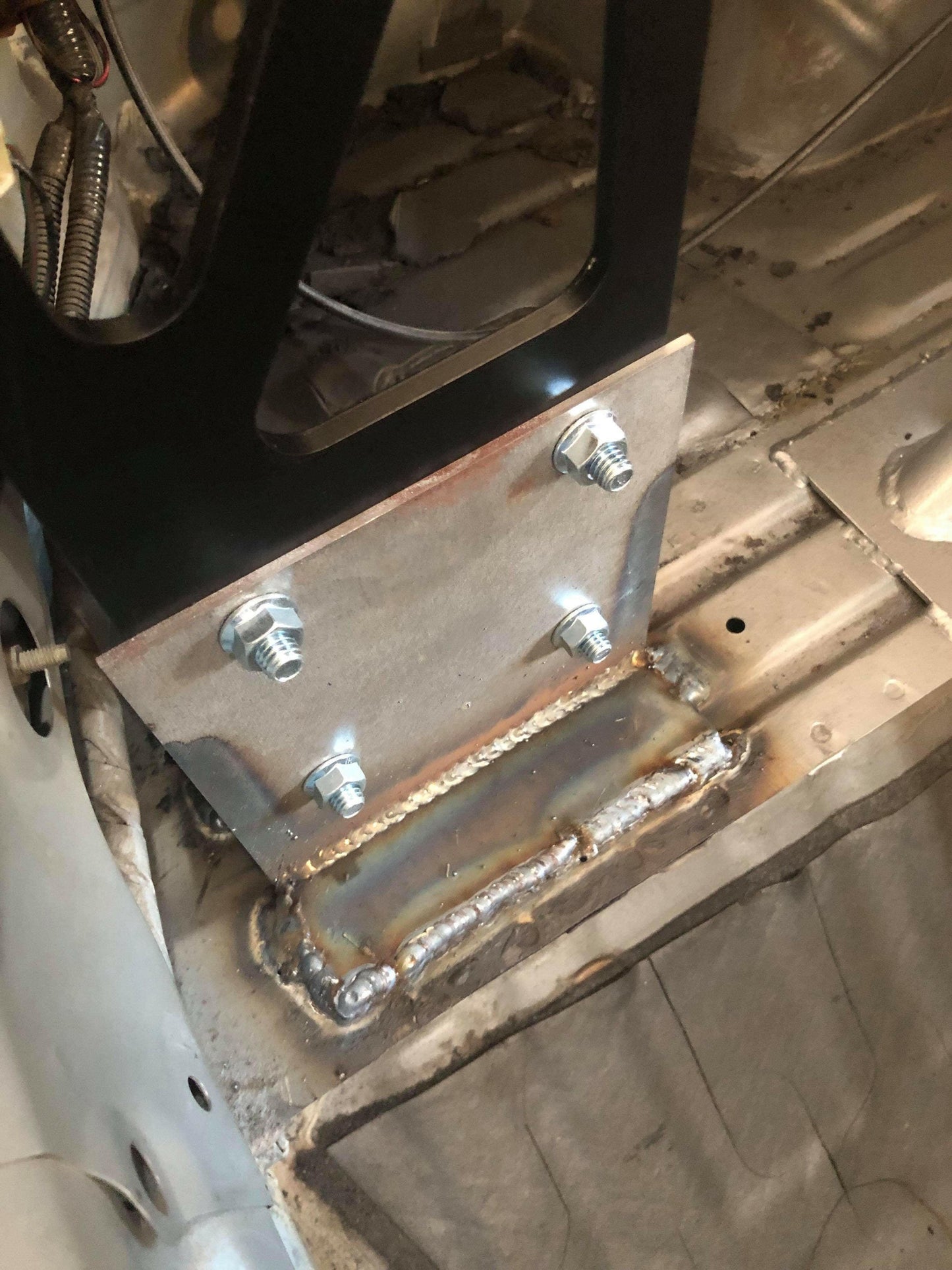 A close-up view of a 9livesracing Custom Chassis Mount kit bolted and welded onto a vehicle's bare floor. The mount is secured with three bolts and features a sturdy weld along its base, seamlessly joining it to the vehicle's chassis. Electrical wires and the vehicle's interior are partially visible.