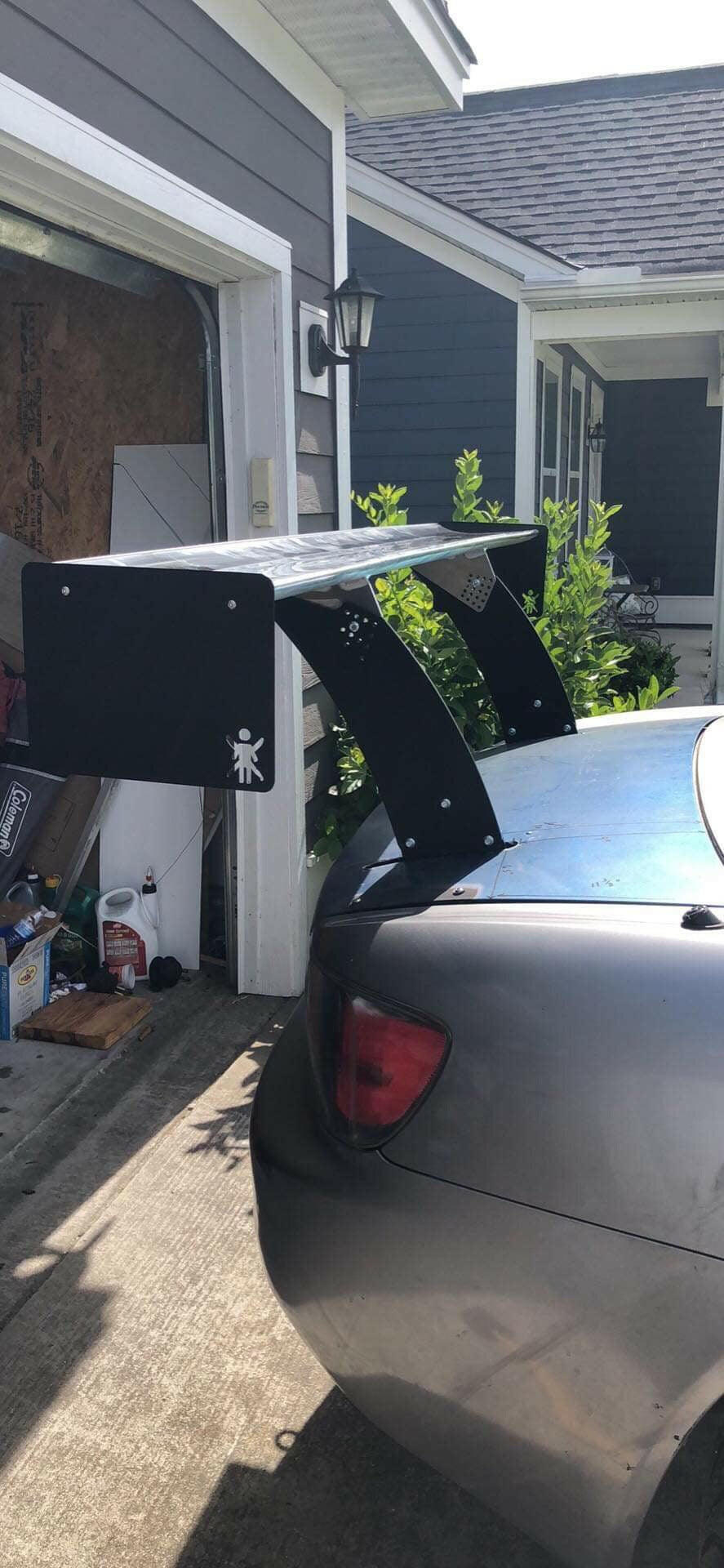 A close-up of a car equipped with 9livesracing's Custom Chassis Mount kits, showcasing a large spoiler with an aggressive wing angle of attack (AOA) attached to its trunk, is parked in front of a house with gray siding. The garage door is open, revealing various items inside. The house has a small outdoor lantern on the wall and greenery next to the driveway.