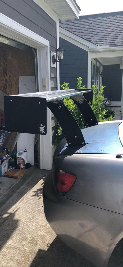 A close-up of a car equipped with 9livesracing's Custom Chassis Mount kits, showcasing a large spoiler with an aggressive wing angle of attack (AOA) attached to its trunk, is parked in front of a house with gray siding. The garage door is open, revealing various items inside. The house has a small outdoor lantern on the wall and greenery next to the driveway.