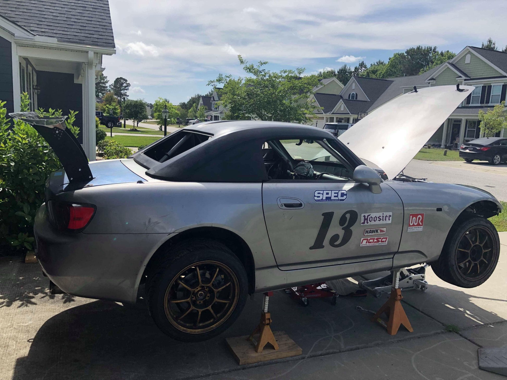 A silver race car, proudly displaying the number 13 along with various sponsor decals, sits elevated on jack stands in a suburban driveway. The car's hood and trunk are open, revealing the intricate 9livesracing Custom Chassis Mount kits. Its right front wheel is removed, with modern houses and lush greenery framing the background.