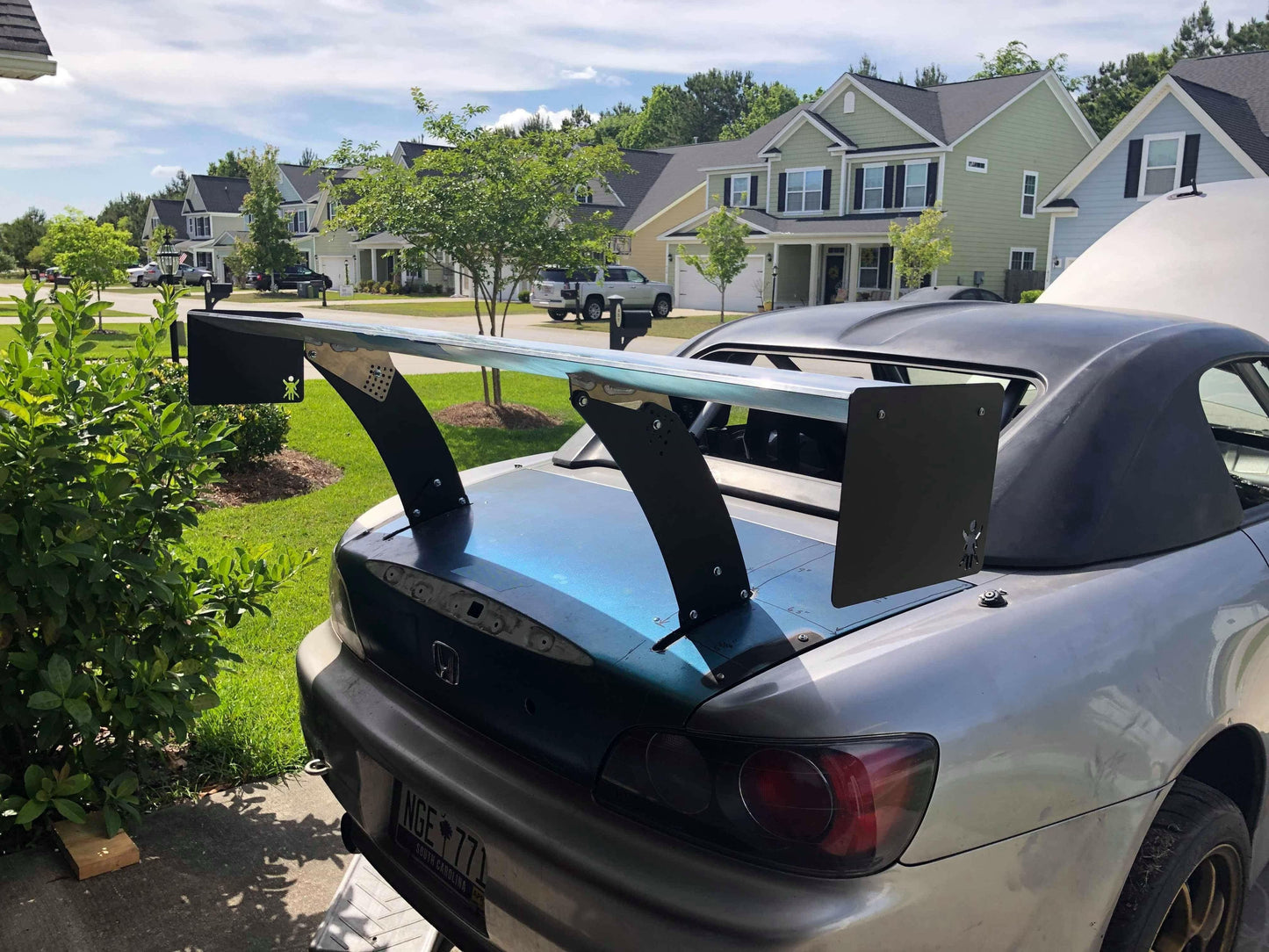 A silver convertible car is parked in a suburban neighborhood driveway, featuring an oversized rear wing spoiler equipped with 9livesracing Custom Chassis Mount kits. Houses, a lawn, trees, and parked cars are visible in the background under a partly cloudy sky.