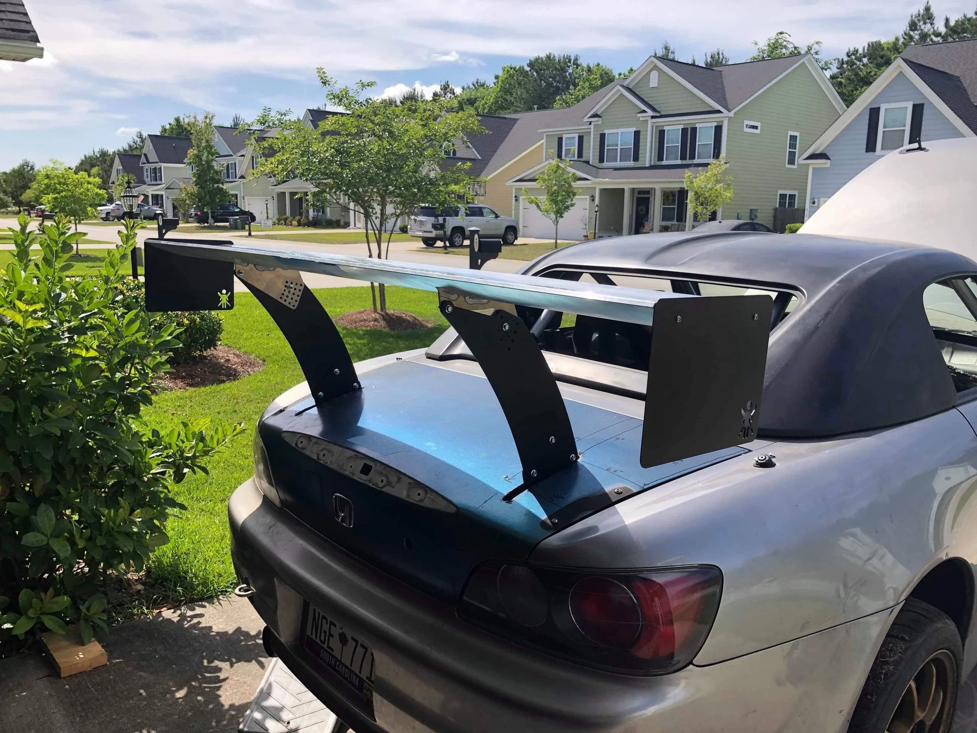 A silver Honda S2000 convertible, equipped with a 9livesracing Chassis Pylons kit and parked in a suburban residential neighborhood, boasts an oversized rear spoiler. The open trunk reveals a neatly maintained environment with green lawns and modern, two-story houses in the background.