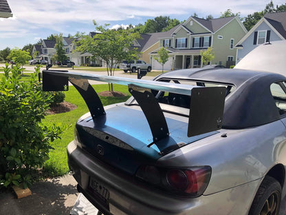 A silver Honda S2000 convertible, equipped with a 9livesracing Chassis Pylons kit and parked in a suburban residential neighborhood, boasts an oversized rear spoiler. The open trunk reveals a neatly maintained environment with green lawns and modern, two-story houses in the background.