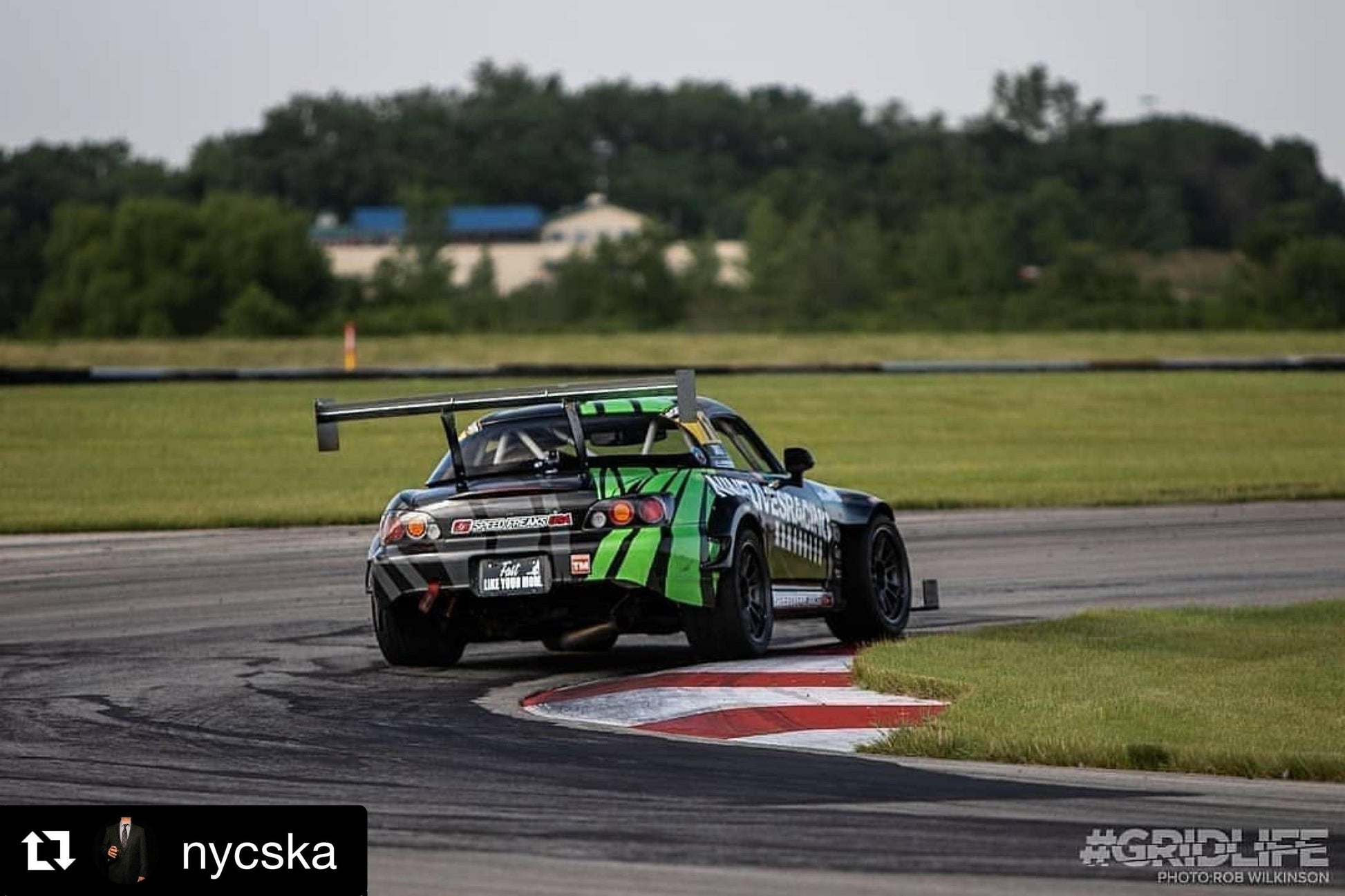 A race car equipped with 9livesracing's Custom Chassis Mount kits, sporting a striking black and green paint job, takes a turn on a racetrack. The impressive chassis mount wing is showcased at an optimal angle of attack (AOA), enhancing the car’s performance. In the background, green grass, trees, and distant buildings can be seen. Photo credit goes to GRIDLIFE, and it was reposted by @nycska.