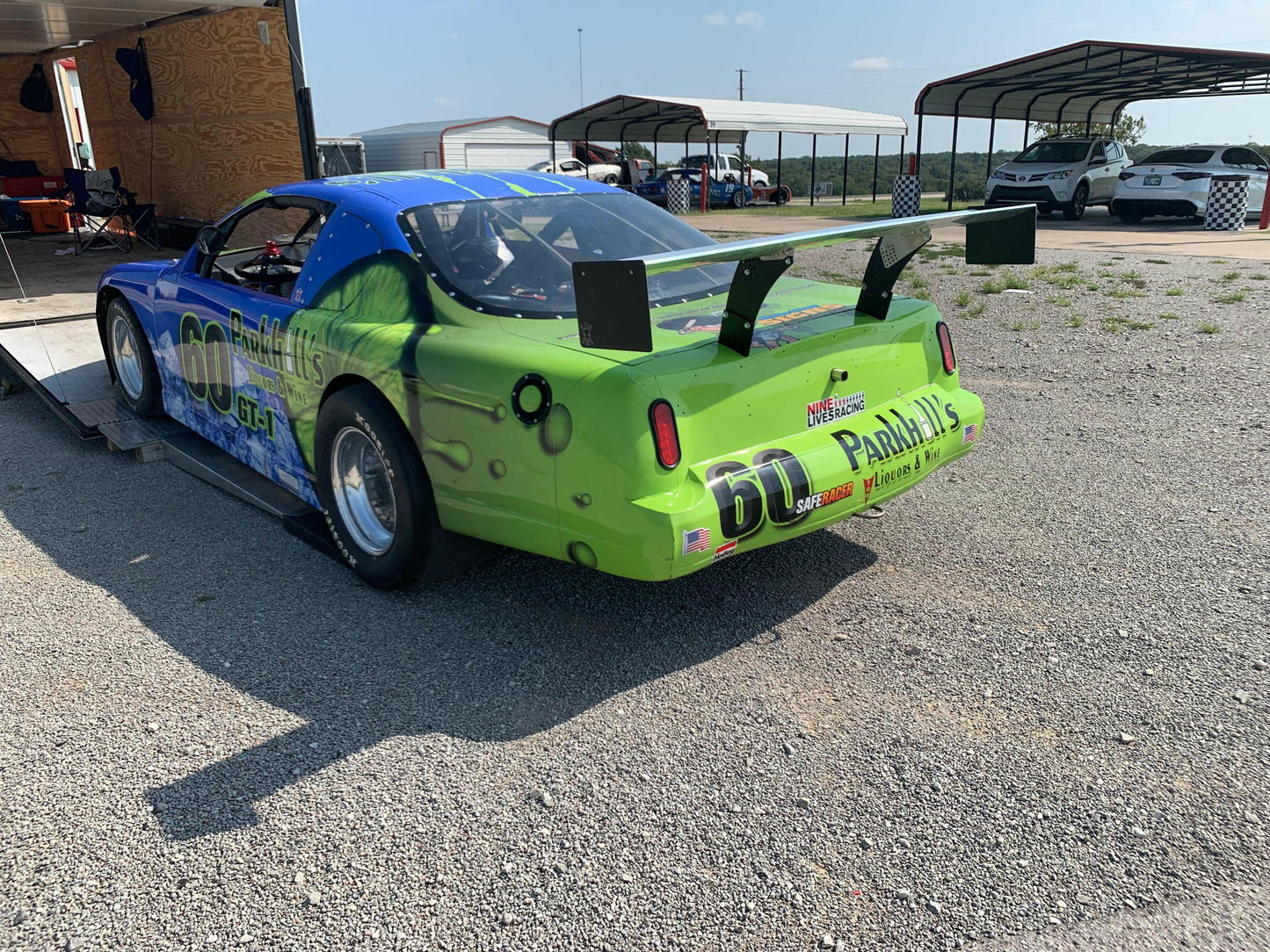 A green and blue racing car featuring a prominent rear wing is parked on a gravel area next to a trailer. The car, numbered "60" with "Parra's" on the back, showcases an optimized Angle Of Attack (AOA) thanks to 9livesracing's Custom Chassis Mount kits. Several other structures and vehicles are visible in the background.