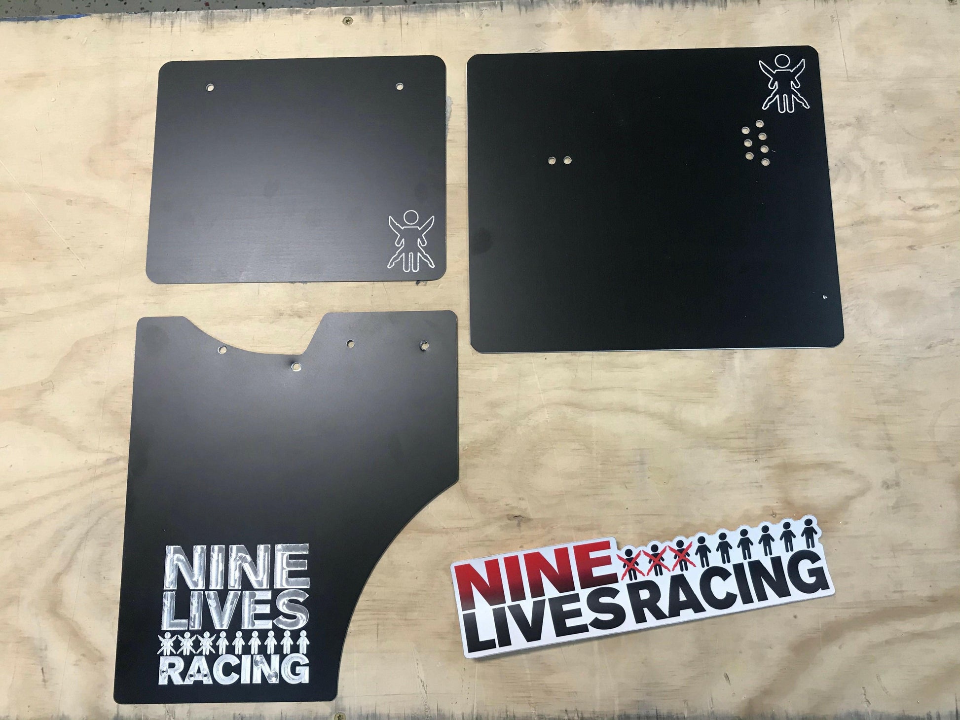 Three black aluminum end plates for The Big Wáng!, possibly car parts, are laid out on a wooden surface bearing the "9livesracing" logo. At the bottom of the image, a sticker with the same "9livesracing" logo and small human-like symbols in a row is also visible.