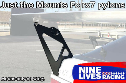 A car is equipped with an aftermarket aluminum Fc Rx7 Wing Mounts - Pylons Only kit by 9livesracing. These mounts are designed to support a spoiler, but the spoiler is not present. The background shows an empty road and a safety barrier. The text reads, "Just the Mounts FC Rx7 pylons" and "9livesracing".