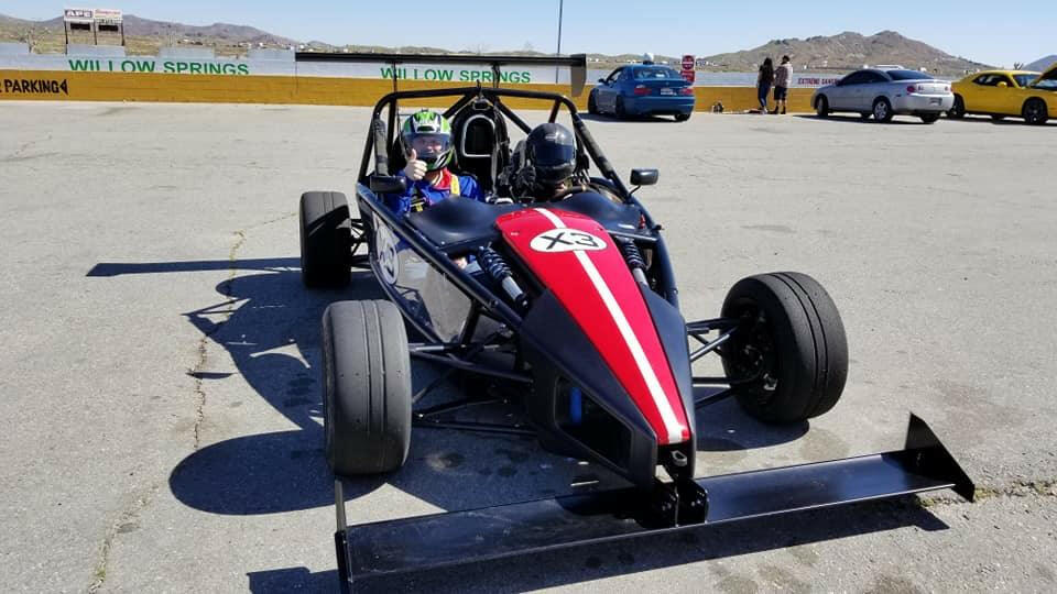 A race car adorned with striking red and white stripes is parked on a concrete lot. Two individuals in racing gear are seated inside, proudly demonstrating the newest 9livesracing Custom Wing Pylons. In the background, a yellow and black "Willow Springs" sign is visible along with more parking area and distant hills.