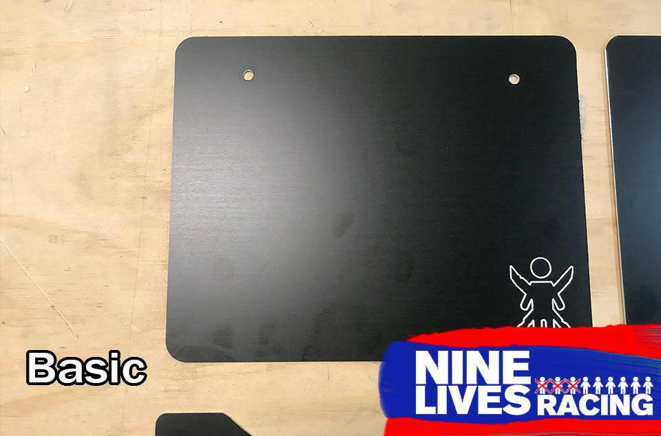 A rectangular black aluminum plate with two holes near the top is displayed against a wooden background. The plate has the word "Basic" on it, and the "9livesracing" logo, renowned for its downforce technology, is partially visible in the bottom right corner.