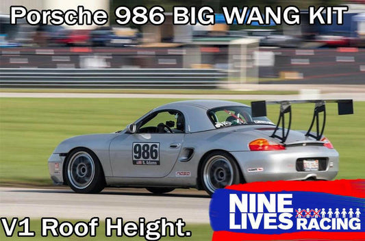 A silver Porsche Boxster 986, outfitted with a prominent rear wing, races on the track. The car sports the number 986 on its door. Overlay text in the image reads, "Porsche Boxster Big Wang Kit '96-04 986 GT3 V1 Roof Height," accompanied by the 9livesracing logo at the bottom, showcasing its exceptional down-force capabilities.