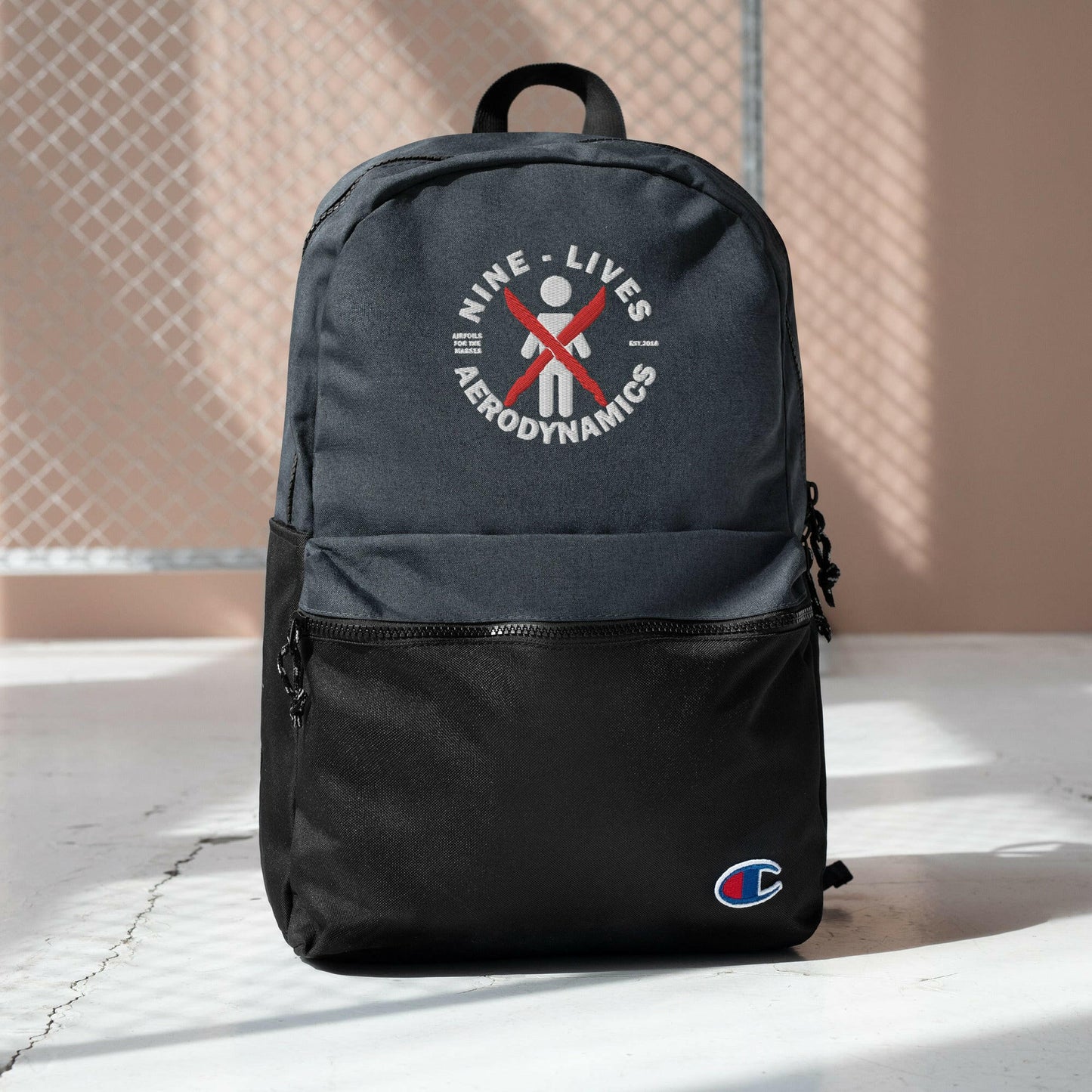 A black and gray water-resistant Embroidered Champion Backpack from Nine Lives Racing, featuring a white and red logo with the stylized figure "NINE LIVES AERODYNAMICS." The small size backpack includes a zippered front pocket and a Champion logo near the bottom right corner. It is displayed against a light-colored background with some shadows.