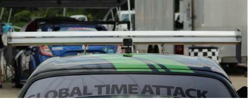 A close-up shot of a sports car's rear wing, showcasing 9livesracing's "The Deuce! Double Element Kit," with "Global Time Attack" emblazoned on the back windshield. The car features striking green and black stripes. Another car and equipment are visible in the background in a paddock area.