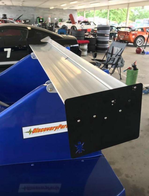 A blue vehicle in the garage sports 9livesracing’s American-made, large metal spoiler, The Deuce! Double Element Kit. The space is filled with tools, equipment, and car parts. In the background, several cars are visible, including one with racing numbers and another orange vehicle.