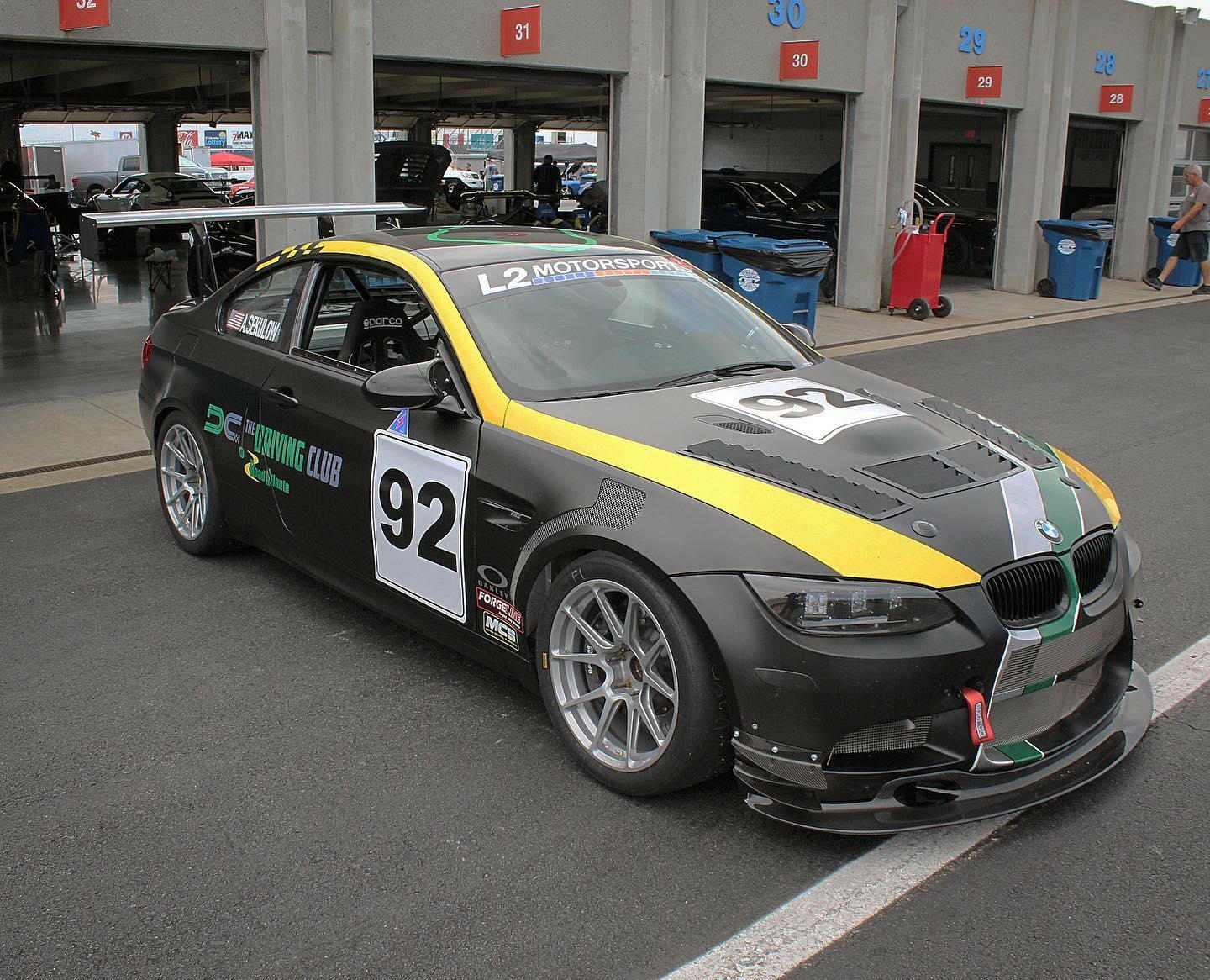 A 3-Series Big Wang kit ’04-13 E90/E92/E93 from 9livesracing, featuring a sleek black design with number 92 and adorned with yellow and green stripes, is parked in a pit garage. This race car boasts extruded aluminum wings for additional downforce, along with various sponsor logos, a spoiler, racing wheels, and aerodynamic enhancements. In the background are several other cars and garage doors numbered 28 to 32.