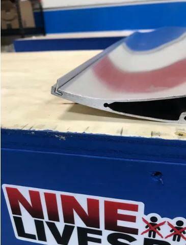 A close-up of a Gurney Flap with a beveled edge, placed on a wooden surface. In the background, there's a blue and white sign with the text "9livesracing" and a small logo of two stick figures.