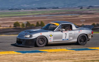 A silver race car with the number 7 on its doors speeds along a racetrack during a race, equipped with a Miata Damn Dam '90-05 NA/NB by 9livesracing for added downforce. The car, possibly inspired by Mazda Miatas, is adorned with various sponsor stickers. The background features fields and hills under a blue sky. Photo by Mike Doran, 2020.