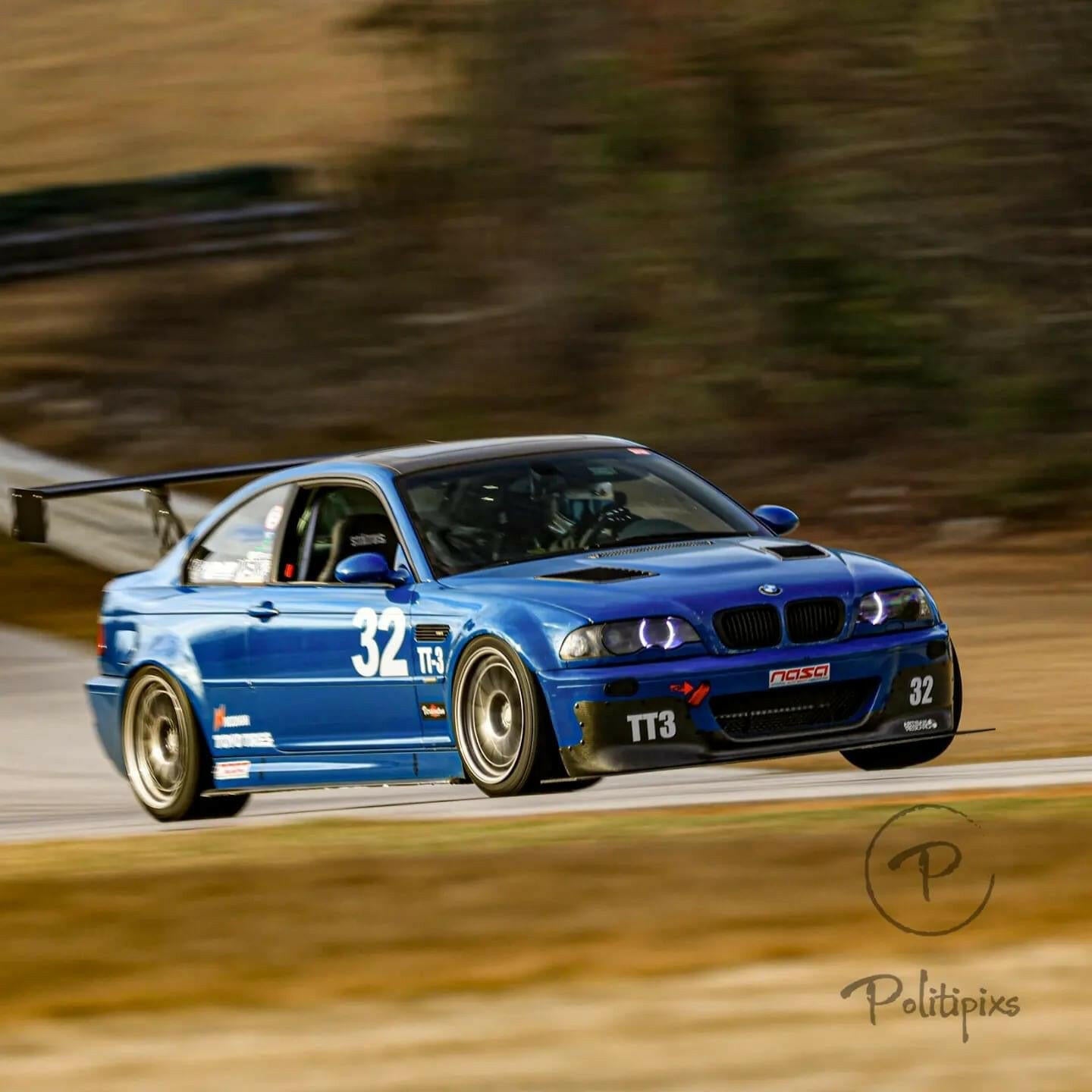 A blue BMW race car from the 3-Series Big Wang Kit '97-06 E46 by 9livesracing, emblazoned with the number '32' and 'TT3' on the doors, is speeding on a racetrack. Featuring a Big Wang GT3 rear wing, the car showcases various decals. The background shows blurred scenery indicating motion, and the image is watermarked with "Politipix".