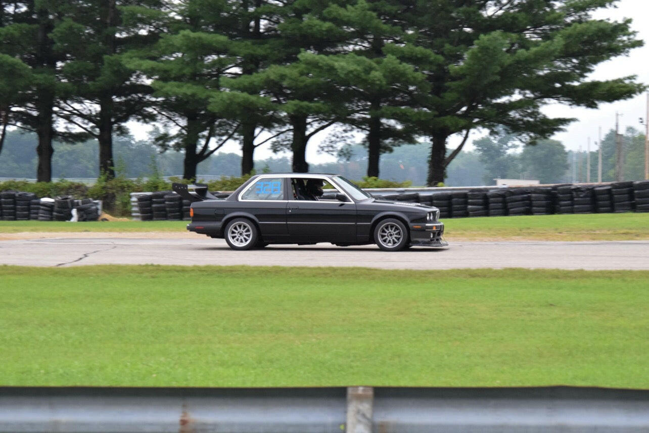 A black race car fitted with a 9livesracing Street Wang for the 82-92 BMW 318 and 325 E30 speeds along a paved track, with a green grassy area and rows of tires in the background. Tall trees line the track, creating a scenic backdrop. The car has tinted windows and showcases impressive aerodynamic performance during the race.