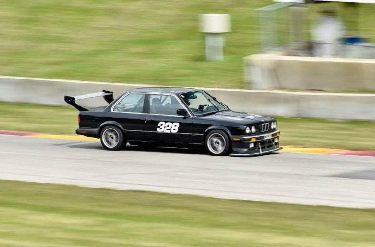 A black Street Wang 82-92 BMW 318 325 E30 with the number 328 on the door speeds down a track lined with grass. Equipped with a large rear spoiler and front splitter for optimal aerodynamic performance, the car's motion blur emphasizes its high speed. This high-performance model from 9livesracing showcases exceptional agility and power.