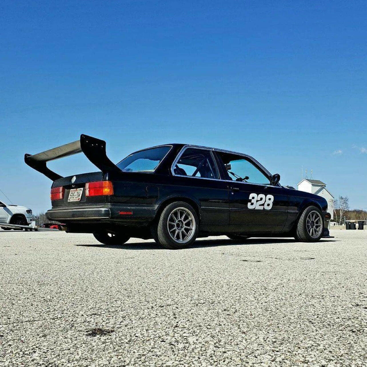 A Street Wang 82-92 BMW 318 325 E30, in sleek black with the number 328 on its side, is parked on an open concrete area under a clear blue sky. This racing car, equipped with a Miata wang kit from 9livesracing, boasts a prominent rear wing and sporty modifications for enhanced aerodynamic performance. In the background, another vehicle and some small structures are visible.