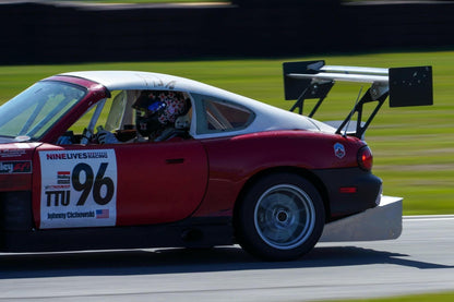 A Miata Diffuser '90-05 NA/NB from 9livesracing, installed on a red and white race car with the number 96 on the door and an aluminum rear wing, is speeding on a track. The V8 Miata displays visible sponsor decals. The driver, partially visible through the window and wearing a helmet, adds to the dynamic scene as the blurred background emphasizes their motion.