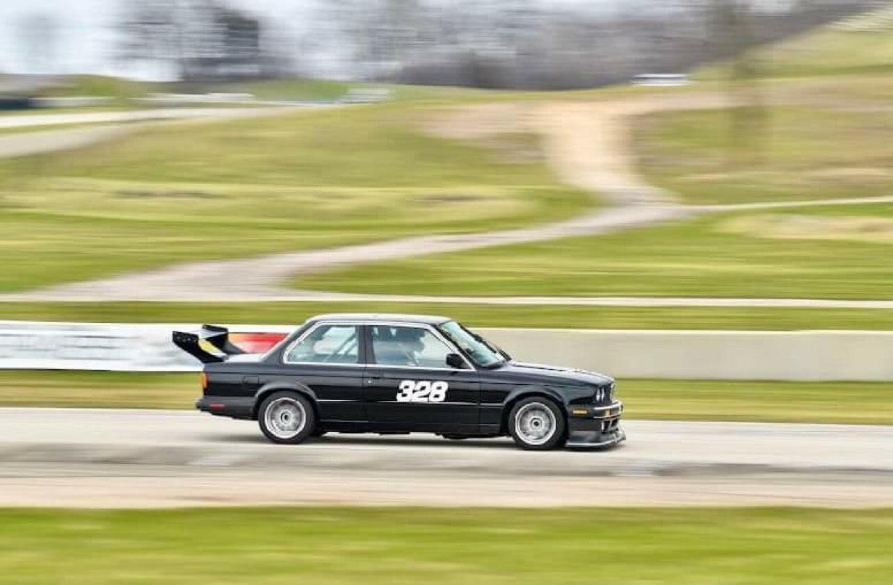 A Street Wang 82-92 BMW 318 325 E30 by 9livesracing, painted black and featuring the number 328 on its side, speeds along the track, showcasing its aerodynamic performance. The blurred backgrounds of grass and a dirt road depict motion as this car, equipped with a large rear wing and low-profile tires, ensures every turn is mastered.