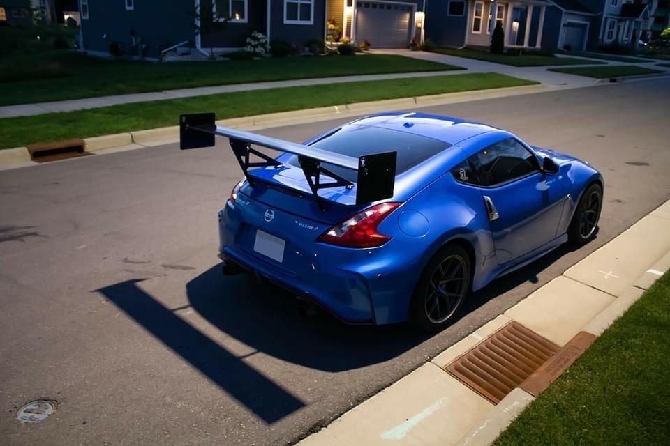 A blue sports car, featuring the 370z Big Wang Kit '08-20 Z34 by 9livesracing, with a large extruded aluminum rear wing, is parked on a residential street. The car, equipped with advanced down-force adjustments, is near the curb, surrounded by houses, driveways, and well-maintained lawns. The street remains quiet and empty.