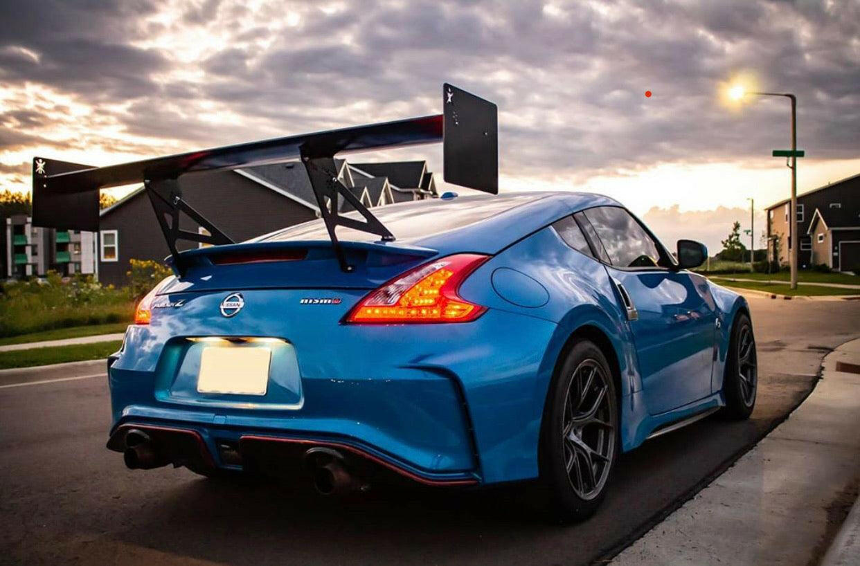 A vivid blue 370z Big Wang Kit '08-20 Z34 from 9livesracing, featuring extruded aluminum wings and a large rear spoiler, is parked on a suburban street at sunset. The car's taillights are illuminated, while dark clouds in the sky are partially lit by the setting sun. Houses and street lights can be seen in the background.